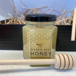 May include: A glass jar of honey with a black lid and a label that reads "Queen Bee Honey Produced in England". A wooden honey dipper is in the foreground.