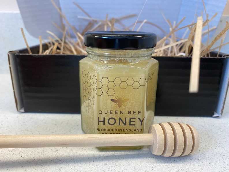 May include: A glass jar of honey with a black lid and a label that reads "Queen Bee Honey Produced in England". A wooden honey dipper is in the foreground.