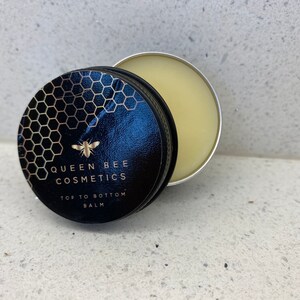 May include: A small, round tin container with a black lid featuring a gold honeycomb pattern, a bee, and the text "Queen Bee Cosmetics Top to Bottom Balm". The tin is open, revealing a pale yellow balm inside.