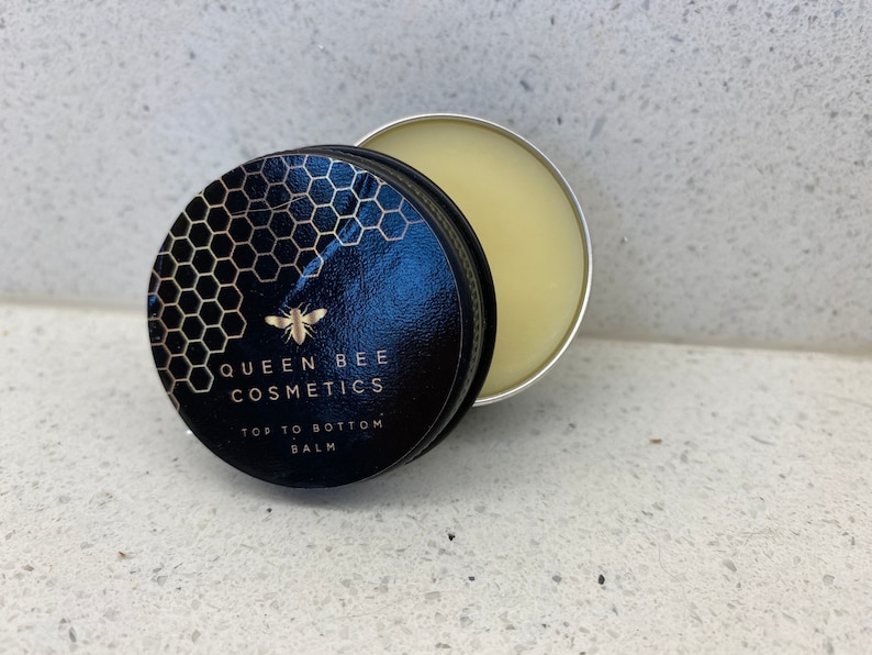 May include: A small, round tin container with a black lid featuring a gold honeycomb pattern, a bee, and the text "Queen Bee Cosmetics Top to Bottom Balm". The tin is open, revealing a pale yellow balm inside.