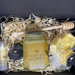 May include: A gift box filled with honey-themed beauty products. The box contains a jar of Queen Bee Honey, a lip balm, a small jar of top to bottom balm, and a bag of honey soap bars. The box is filled with wood shavings and a wooden honey dipper.