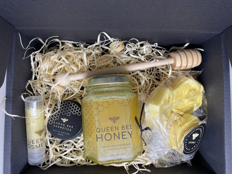 May include: A gift box filled with honey-themed beauty products. The box contains a jar of Queen Bee Honey, a lip balm, a small jar of top to bottom balm, and a bag of honey soap bars. The box is filled with wood shavings and a wooden honey dipper.