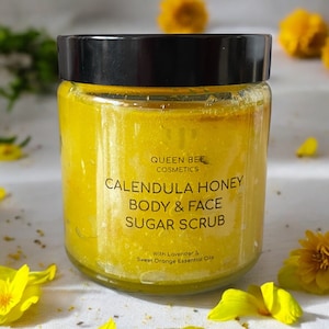 May include: A glass jar of yellow calendula honey body and face sugar scrub with a black lid. The label reads "Queen Bee Cosmetics Calendula Honey Body & Face Sugar Scrub With Lavender & Sweet Orange Essential Oils".