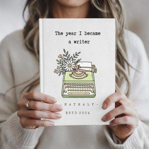 May include: A white hardcover journal featuring a light green typewriter illustration adorned with a bouquet of flowers. The text reads: The year I became a writer, NATHALY, and ESTD 2026. The design uses black, green, yellow, and orange ink.