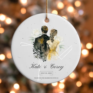 Puede incluir: Un adorno redondo de cerámica blanca con una ilustración en acuarela de una pareja vista desde atrás, con el texto Kate & Corey, July 22, 2023, y Thank you for celebrating with us. El diseño incluye flores blancas y un cordón dorado para colgar.