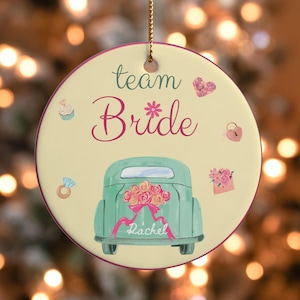 Puede incluir: Un adorno redondo de cerámica con el texto team Bride en letra cursiva verde oscuro y rosa. El diseño incluye un coche vintage de color verde claro con un ramo de flores, un anillo de diamantes, un cupcake, un candado en forma de corazón y un sobre.
