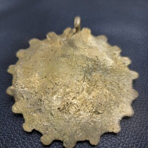 May include: A gold-colored metal pendant with a scalloped edge. The pendant has a loop at the top for hanging.