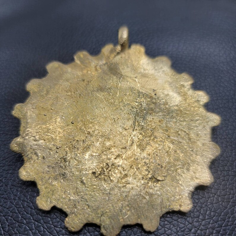 May include: A gold-colored metal pendant with a scalloped edge. The pendant has a loop at the top for hanging.