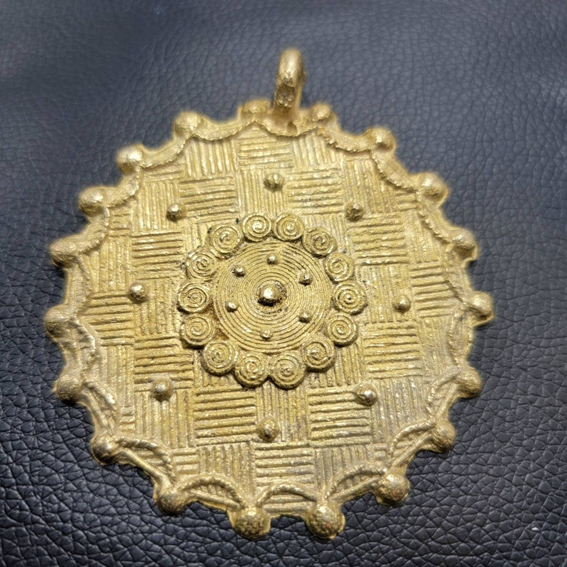 May include: A gold-colored pendant with a circular design. The pendant has a central circle with a spiral pattern surrounded by a grid pattern. The pendant has a loop at the top for attaching to a chain.