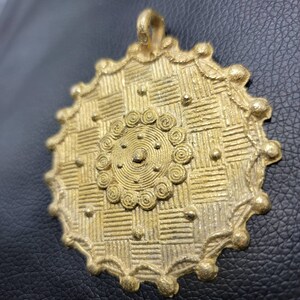 May include: A gold-colored metal pendant with a circular design. The pendant features a central circular motif with concentric circles and a raised dot in the center. The outer edge of the pendant is decorated with a series of small, raised dots.