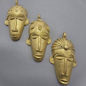 May include: Three gold-colored metal pendants in the shape of stylized human faces. Each pendant has a different design and features intricate details.
