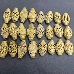 May include: A collection of twenty-one gold-colored, elongated beads. Each bead features unique decorative patterns, including geometric cutouts, spiral designs, and cross-like shapes. The beads are arranged on a dark surface, showcasing their intricate details.
