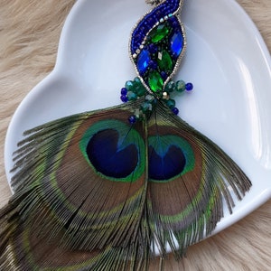 Puede incluir: Un broche de pavo real con cuentas y plumas iridiscentes azules y verdes. El broche est&aacute; en un plato blanco en forma de coraz&oacute;n.