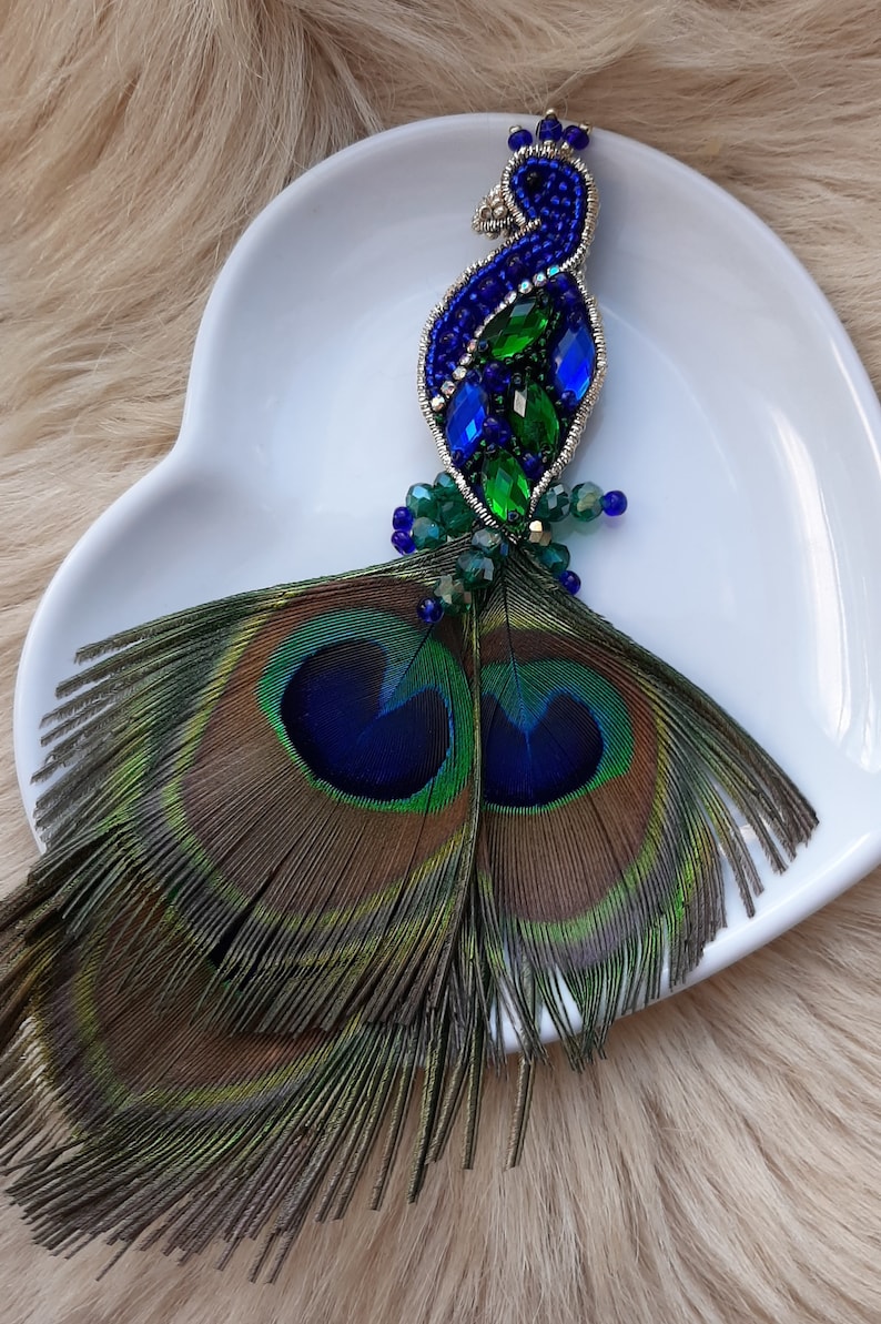 Puede incluir: Un broche de pavo real con cuentas y plumas iridiscentes azules y verdes. El broche est&aacute; en un plato blanco en forma de coraz&oacute;n.