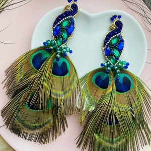 Puede incluir: Un par de pendientes de plumas de pavo real con pavos reales de cuentas azules y verdes. Los pendientes miden aproximadamente 15 cm de largo.