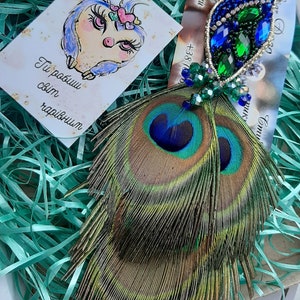 Puede incluir: Un par de pendientes de plumas de pavo real con adornos de cristal azul y verde. Los pendientes se muestran en una caja con papel verde triturado.