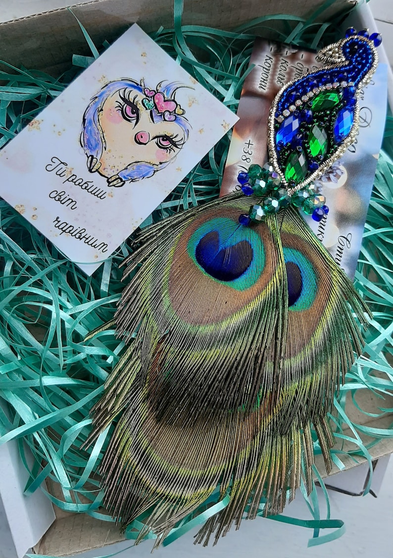 Puede incluir: Un par de pendientes de plumas de pavo real con adornos de cristal azul y verde. Los pendientes se muestran en una caja con papel verde triturado.
