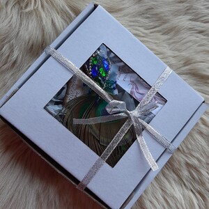 Puede incluir: Una caja de regalo blanca con una cinta plateada atada en un lazo. La caja contiene un broche de pluma de pavo real con detalles en verde, azul y dorado. El broche es visible a trav&eacute;s de una ventana transparente en la caja.