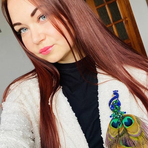 Puede incluir: Una mujer con el pelo largo y rojo oscuro lleva un c&aacute;rdigan blanco y un jersey de cuello alto negro. Lleva un broche con tem&aacute;tica de pavo real con plumas azules, verdes y doradas.