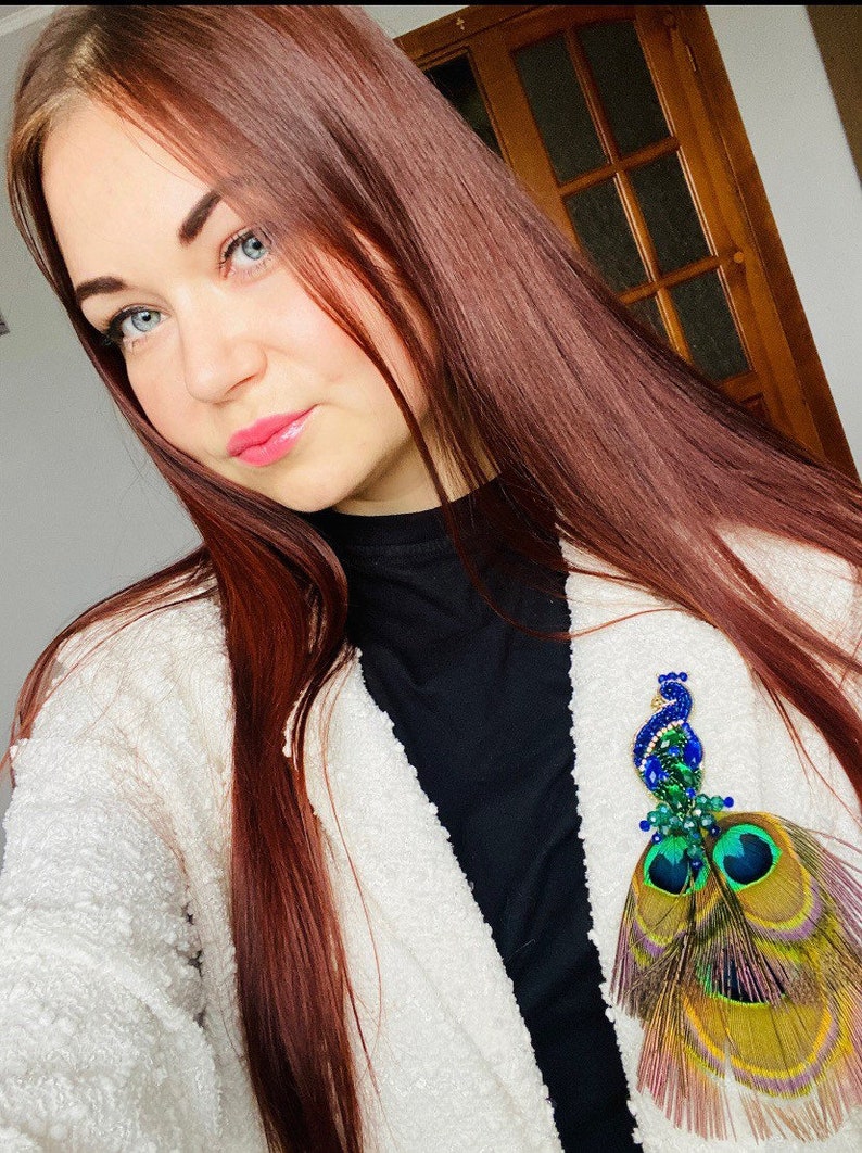Puede incluir: Una mujer con el pelo largo y rojo oscuro lleva un c&aacute;rdigan blanco y un jersey de cuello alto negro. Lleva un broche con tem&aacute;tica de pavo real con plumas azules, verdes y doradas.
