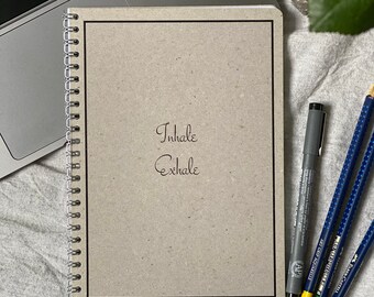May include: A brown notebook with a black border and a silver spiral binding. The notebook has the words "Inhale" and "Exhale" written on the front in black ink.