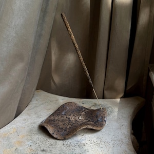 May include: A brown and black speckled ceramic incense holder with an irregular, wavy edge shape. A single, thin, textured brown incense stick is inserted into the holder, standing upright at an angle. The surface of the holder has a glossy finish.