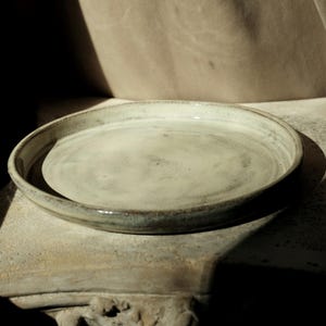 May include: A round ceramic plate with a low, raised rim. The surface features a mottled, off-white glaze with subtle grey and brown speckles throughout. The edges of the plate show a darker, earthy tone where the glaze thins, revealing the clay.