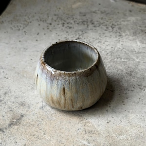 May include: A small, round ceramic bowl with a wide opening and a tapered base. The exterior features a reactive glaze with vertical streaks of beige, tan, and muted gray tones, while the rim is finished with a darker, textured brown edge.