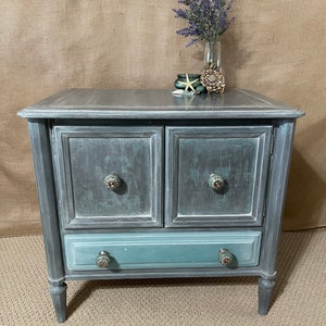 May include: A gray and blue painted wooden end table with two doors and a drawer. The table has decorative knobs and sits on four turned legs.