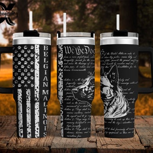 May include: A black stainless steel travel mug with a handle and a black lid. The mug features a design of a Belgian Malinois dog with an American flag background. The text "We the Dog" is printed on the mug, along with a portion of the US Constitution.
