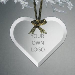 Puede incluir: Un adorno de vidrio transparente en forma de corazón con el texto "YOUR OWN LOGO" grabado en la parte delantera. El adorno cuelga de una cinta y un lazo dorados. El fondo es un degradado de gris.