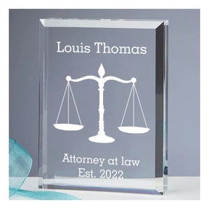 May include: A clear glass block with the name "Louis Thomas" and the scales of justice symbol etched in white. The text "Attorney at law Est. 2022" is also displayed. The block is rectangular and sits on a white surface.