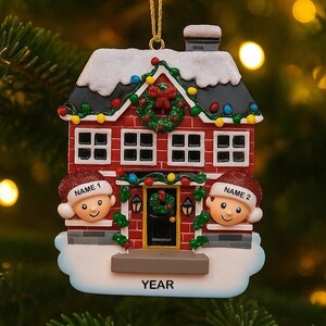 May include: A red and white Christmas house ornament with two smiling faces wearing Santa hats. The house has a wreath on the door and is decorated with lights and snow. The word "YEAR" is at the bottom.