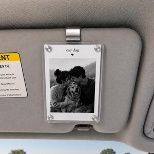 May include: A clear acrylic car visor photo frame featuring a black and white photograph of two people and a dog. The frame is secured with a silver metal clip and includes the printed text our dog with a small heart icon above the photo.