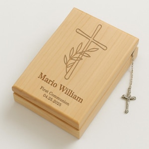 May include: A wooden box with a cross and olive branch design engraved on the lid. The box is personalized with the name "Mario William", the text "First Communion", and the date "04.25.2025". A silver chain with a cross charm hangs from the box.