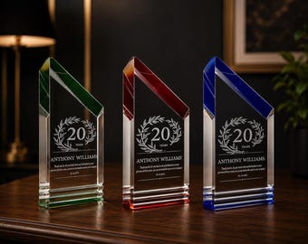 May include: Three glass awards with colored tops in green, red, and blue. Each award has a clear body with the text "20 YEARS" inside a laurel wreath, along with a name and a message. The awards are displayed on a wooden surface.