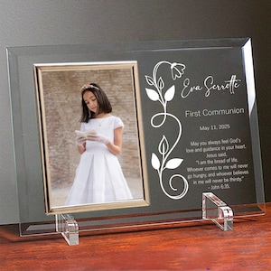 May include: A glass photo frame with a white floral design and the text "Eva Serrette First Communion May 11, 2025". The frame has a photo of a girl in a white dress holding a book.