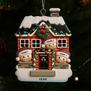 May include: A festive Christmas ornament shaped like a red brick house with three smiling faces wearing Santa hats. The house is decorated with a wreath, lights, and snow. Each face has a name label, and the base reads "YEAR".