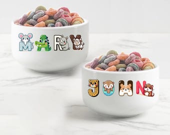May include: Two white ceramic bowls with colorful cereal inside. The bowls have cartoon animal letters spelling out the names "MARY" and "JOHN".