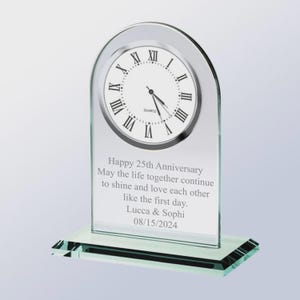 May include: A clear glass anniversary clock with a quartz movement. The clock face has black Roman numerals. The base of the clock has a rectangular shape. The clock is engraved with a 25th anniversary message, names, and a date.