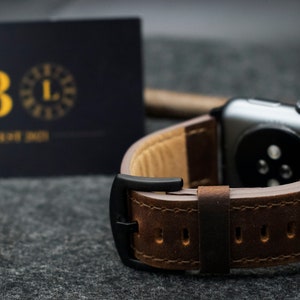 May include: A dark brown leather watch strap with visible stitching and a matte black buckle attached to a dark grey smartwatch case. In the background, a dark rectangular card displays the gold letter B and the text EST 2021.