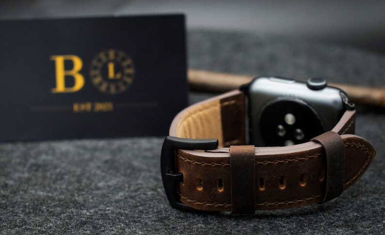 May include: A dark brown leather watch strap with visible stitching and a matte black buckle attached to a dark grey smartwatch case. In the background, a dark rectangular card displays the gold letter B and the text EST 2021.