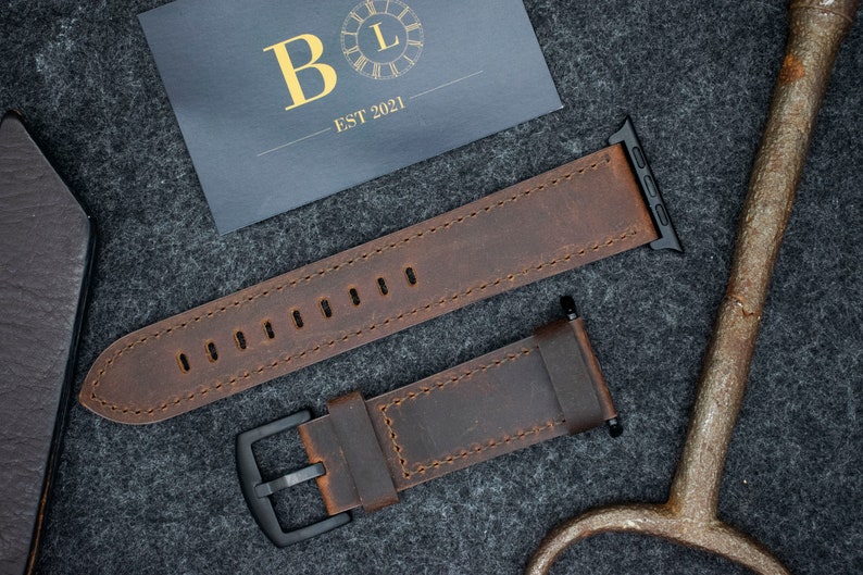 May include: A brown leather watch strap featuring visible stitching, a black metal buckle, and black attachment hardware. A dark rectangular card is present with the gold text B L and EST 2021, alongside a metallic tool with a rounded handle.