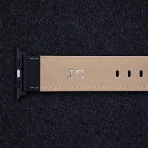 May include: A watch strap featuring a black leather end with a silver-toned connector and a beige leather strap section. The beige section is embossed with the silver-coloured capital letters JC and includes three rectangular adjustment holes.