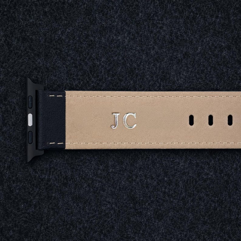May include: A watch strap featuring a black leather end with a silver-toned connector and a beige leather strap section. The beige section is embossed with the silver-coloured capital letters JC and includes three rectangular adjustment holes.
