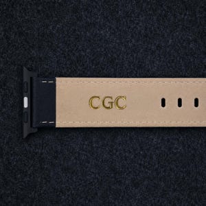 May include: A black watch strap with a beige interior lining featuring the gold embossed letters CGC. The strap includes a black connector piece on the left side and three visible adjustment holes on the right side with white stitching.