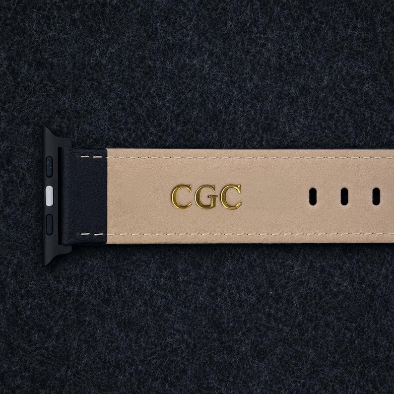 May include: A black watch strap with a beige interior lining featuring the gold embossed letters CGC. The strap includes a black connector piece on the left side and three visible adjustment holes on the right side with white stitching.