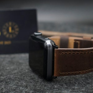 May include: A dark grey smartwatch with a rectangular screen and a brown leather strap featuring visible stitching. The watch is positioned next to a dark rectangular card with the gold letter B and the text EST 2021 printed on the front surface.