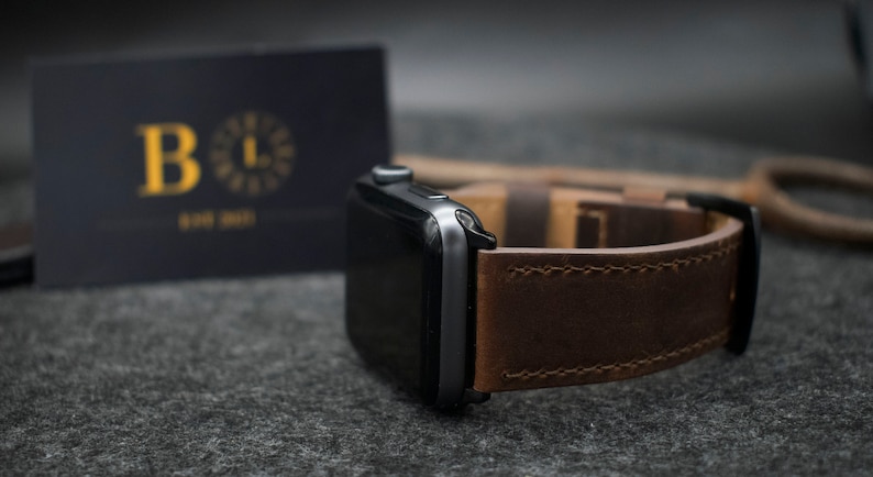 May include: A dark grey smartwatch with a rectangular screen and a brown leather strap featuring visible stitching. The watch is positioned next to a dark rectangular card with the gold letter B and the text EST 2021 printed on the front surface.