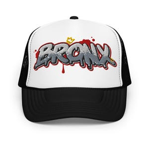 Puede incluir: Una gorra de camionero negra y blanca con un diseño de graffiti de estilo "BRONX" rojo y blanco con una corona dorada en la parte superior.